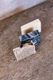 Three blocks of artisanal soap are arranged on a textured surface. The soaps vary in color and texture; one has a white base with black flecks, another is creamy with a smooth finish, and the third has a marble-like, swirled pattern.