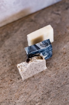 Three blocks of artisanal soap are arranged on a textured surface. The soaps vary in color and texture; one has a white base with black flecks, another is creamy with a smooth finish, and the third has a marble-like, swirled pattern.