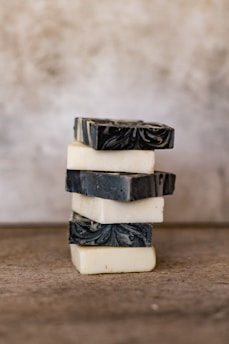 A stack of artisan soap bars is arranged on a neutral brown surface. The bars alternate between creamy white and deep gray colors, with the darker bars featuring an intricate marbled pattern. The background is softly blurred, adding a rustic and minimalistic feel.
