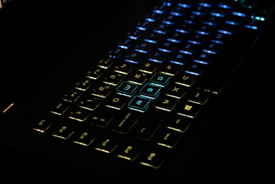 A close-up view of a computer keyboard with illuminated, multicolored backlit keys predominantly featuring shades of blue and green. The highlighted WASD keys stand out in blue and are commonly used for gaming.