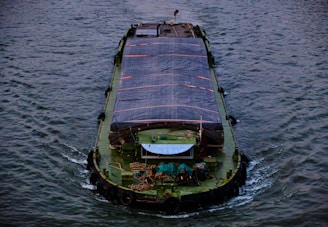 A large cargo barge is moving through a body of water with a covered deck area. The water appears textured with ripples, and the barge has various equipment and ropes on its deck.
