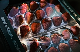 A package of strawberries neatly arranged in a plastic container, with a clear overlay that reflects light. The berries appear ripe and vibrant under soft lighting, with text visible on the package.