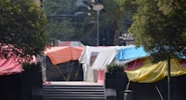 A group of colorful tents are set up on a paved area between trees. The tents are made of tarpaulin in various colors including orange, blue, red, white, black, and yellow. There are steps leading up to the tents, with some greenery visible around the area.
