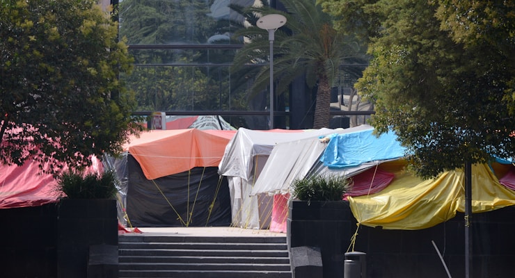 A group of colorful tents are set up on a paved area between trees. The tents are made of tarpaulin in various colors including orange, blue, red, white, black, and yellow. There are steps leading up to the tents, with some greenery visible around the area.