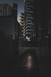 A dark urban scene featuring tall, modern buildings arranged closely together. The architecture includes geometric shapes and sharp edges with a mix of glass and concrete surfaces. There is a subtle light source, perhaps from the setting sun, creating contrast with shadows on the ground and buildings.