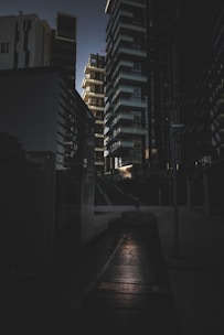 A dark urban scene featuring tall, modern buildings arranged closely together. The architecture includes geometric shapes and sharp edges with a mix of glass and concrete surfaces. There is a subtle light source, perhaps from the setting sun, creating contrast with shadows on the ground and buildings.