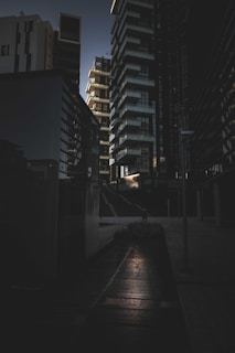 A dark urban scene featuring tall, modern buildings arranged closely together. The architecture includes geometric shapes and sharp edges with a mix of glass and concrete surfaces. There is a subtle light source, perhaps from the setting sun, creating contrast with shadows on the ground and buildings.