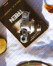 Close-up of a vintage camera resting on a textured notebook beside scattered leaves.