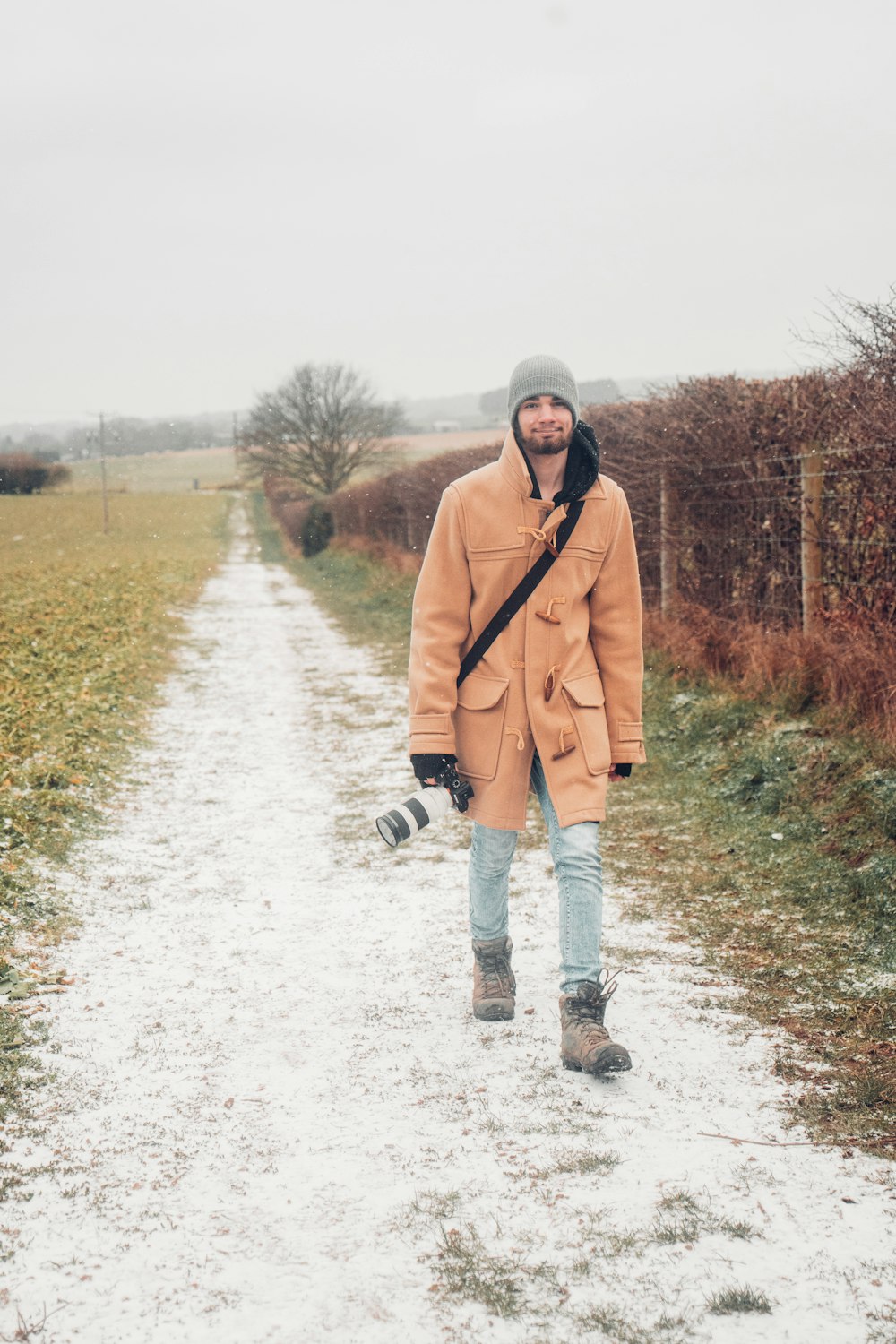 Mann in braunem Mantel und schwarzer Hose geht tagsüber auf schneebedecktem Boden spazieren