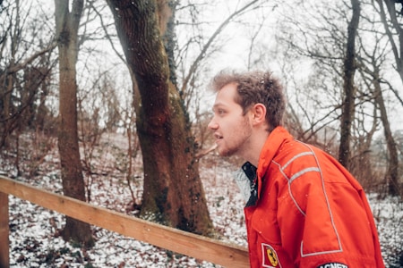 A man wearing a bright orange jacket, embroidered with a logo, is captured outdoors during a snowy day. He is in a woodland area with leafless trees and a light dusting of snow covering the ground. The sky appears overcast, contributing to a cold, wintry atmosphere.