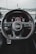A driver's perspective of a car interior focused on the steering wheel with the Audi logo prominently displayed. The dashboard is visible behind the wheel, showing various controls and displays. The stitching on the steering wheel is red, and the overall design is sleek and modern.