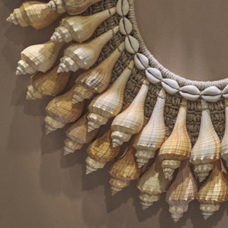 A close-up view of a decorative piece featuring a circular arrangement of seashells. The shells are intricately attached to a woven or knitted base, with alternating small cowrie shells and pointed conch shells creating an ornate pattern. The composition appears artfully crafted, emphasizing natural textures and organic shapes.