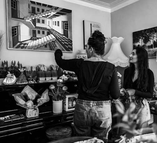 Two people are indoors, engaged in a conversation. One is pointing at a painting of a complex architectural design featuring multiple staircases. The room is decorated with various items such as vases, teapots, and small sculptures, suggesting an artistic or personal space. A large lamp stands nearby, adding an element of coziness.