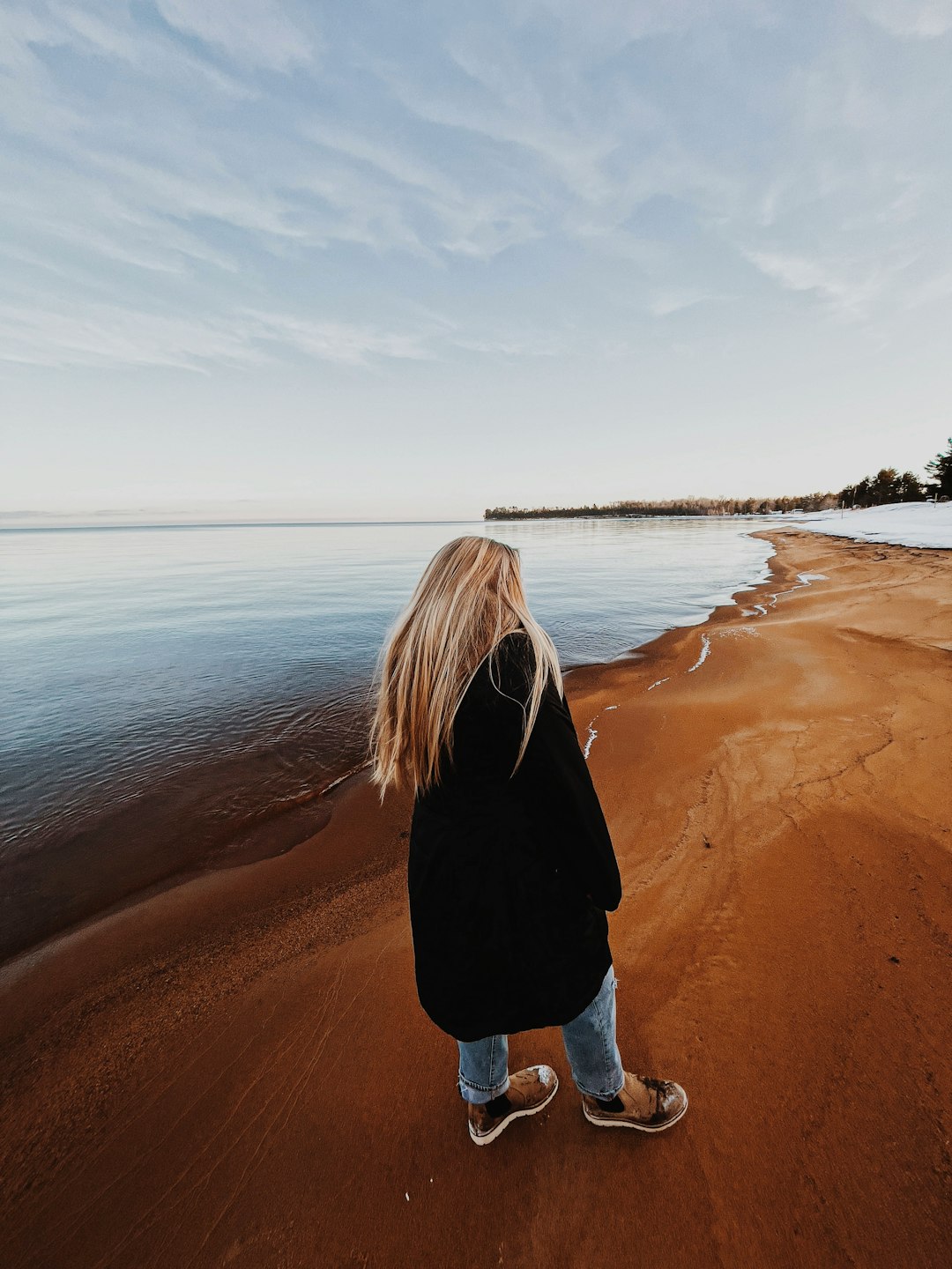 woman in black jacket and blue denim jeans standing on brown sand near body of water