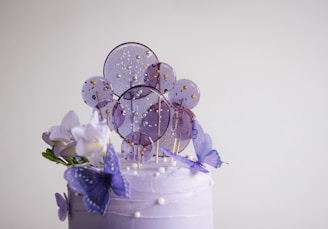 A decorated cake topped with purple and white frosting, adorned with sugar lollipops, purple butterflies, and white flowers. The lollipops are circular with golden beads, adding a sparkling effect.