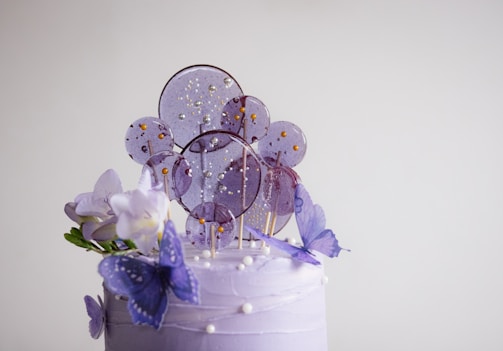 A decorated cake topped with purple and white frosting, adorned with sugar lollipops, purple butterflies, and white flowers. The lollipops are circular with golden beads, adding a sparkling effect.