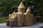 A detailed miniature model of a historic building with intricate architectural features, including stone-like texture, arches, and conical roofs. The model is set outdoors, surrounded by lush green foliage that provides a natural backdrop.