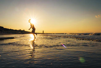 man in white shirt and black shorts jumping on beach during sunset