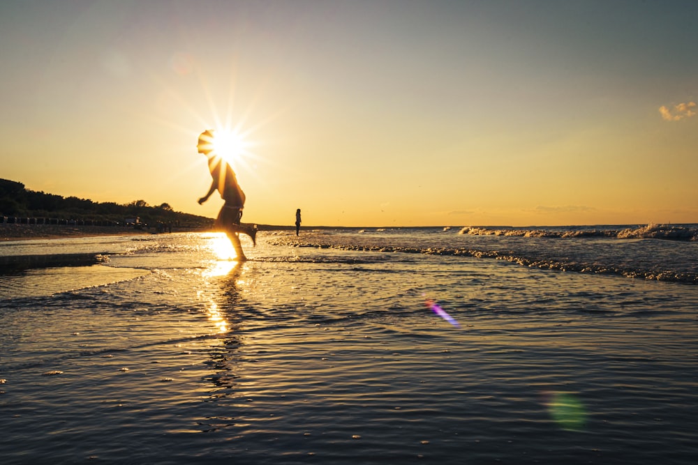 Hombre con camisa blanca y pantalones cortos negros saltando en la playa durante la puesta del sol