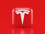 A glossy, red 3D square icon features a white, stylized 'T' logo with a futuristic and sleek design. The background is also solid red, creating a visually striking contrast.