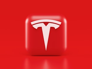 A glossy, red 3D square icon features a white, stylized 'T' logo with a futuristic and sleek design. The background is also solid red, creating a visually striking contrast.