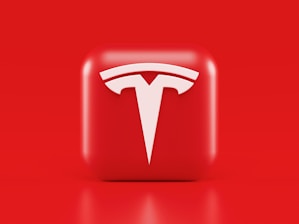 A glossy, red 3D square icon features a white, stylized 'T' logo with a futuristic and sleek design. The background is also solid red, creating a visually striking contrast.