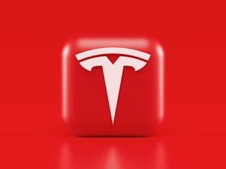 A glossy, red 3D square icon features a white, stylized 'T' logo with a futuristic and sleek design. The background is also solid red, creating a visually striking contrast.