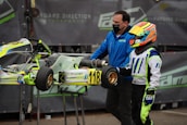 A person wearing a colorful racing suit and helmet stands next to a small racing go-kart with the number 118 displayed. Another individual, wearing a blue jacket and face mask, is assisting with the kart. The background features a motorsport-related banner.