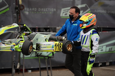 A person wearing a colorful racing suit and helmet stands next to a small racing go-kart with the number 118 displayed. Another individual, wearing a blue jacket and face mask, is assisting with the kart. The background features a motorsport-related banner.
