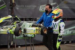 A person wearing a colorful racing suit and helmet stands next to a small racing go-kart with the number 118 displayed. Another individual, wearing a blue jacket and face mask, is assisting with the kart. The background features a motorsport-related banner.