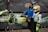 A person wearing a colorful racing suit and helmet stands next to a small racing go-kart with the number 118 displayed. Another individual, wearing a blue jacket and face mask, is assisting with the kart. The background features a motorsport-related banner.