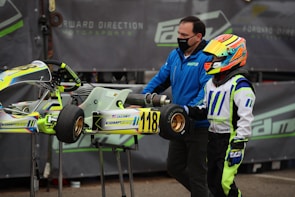 A person wearing a colorful racing suit and helmet stands next to a small racing go-kart with the number 118 displayed. Another individual, wearing a blue jacket and face mask, is assisting with the kart. The background features a motorsport-related banner.