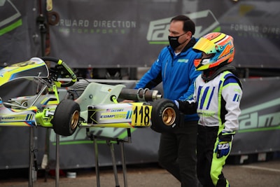 A person wearing a colorful racing suit and helmet stands next to a small racing go-kart with the number 118 displayed. Another individual, wearing a blue jacket and face mask, is assisting with the kart. The background features a motorsport-related banner.