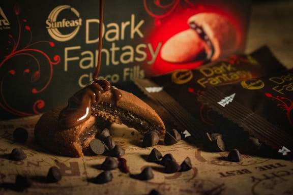 A close-up view of a partially eaten chocolate-filled cookie with melted chocolate dripping from it, surrounded by scattered chocolate chips. The background features a product box and packaging with the label 'Dark Fantasy' displayed prominently, hinting at a rich, indulgent theme.
