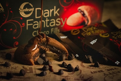 A close-up view of a partially eaten chocolate-filled cookie with melted chocolate dripping from it, surrounded by scattered chocolate chips. The background features a product box and packaging with the label 'Dark Fantasy' displayed prominently, hinting at a rich, indulgent theme.
