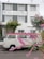 A vintage white van with a red roof is parked in front of a charming hotel named 'Hotel Autre Mer'. The building features green shutters and is surrounded by lush greenery and flowering plants. Pink flowers in the foreground add a touch of color and depth to the scene.