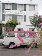 A vintage white van with a red roof is parked in front of a charming hotel named 'Hotel Autre Mer'. The building features green shutters and is surrounded by lush greenery and flowering plants. Pink flowers in the foreground add a touch of color and depth to the scene.