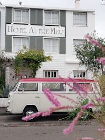 A vintage white van with a red roof is parked in front of a charming hotel named 'Hotel Autre Mer'. The building features green shutters and is surrounded by lush greenery and flowering plants. Pink flowers in the foreground add a touch of color and depth to the scene.