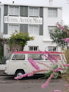 A vintage white van with a red roof is parked in front of a charming hotel named 'Hotel Autre Mer'. The building features green shutters and is surrounded by lush greenery and flowering plants. Pink flowers in the foreground add a touch of color and depth to the scene.