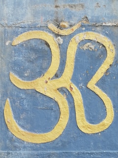 A large yellow Om symbol is painted on a weathered blue concrete wall with noticeable cracks and peeling paint.