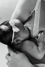A black and white image of an infant breastfeeding. The baby is wearing a delicate headband and is held gently. The adult's arm is visible, cradling the child in a nurturing manner.