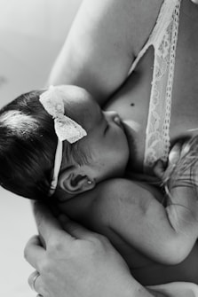 A black and white image of an infant breastfeeding. The baby is wearing a delicate headband and is held gently. The adult's arm is visible, cradling the child in a nurturing manner.