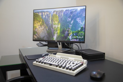 A modern workspace featuring a Dell monitor displaying an image of a lush green canyon. The desk is organized with a vintage-style keyboard, a black mouse, and a leather-bound notebook. The setting appears minimalistic and professional.