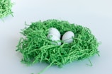 Three white eggs nestled in a bed of green shredded paper, arranged to resemble a nest. The setting is minimalist with a clean white background.