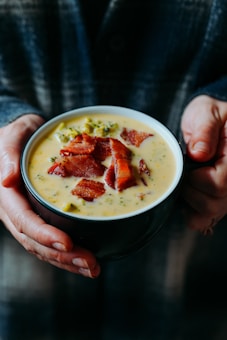 A pair of hands holding a dark bowl filled with creamy soup topped with pieces of crispy bacon and bits of broccoli. The background is softly blurred, suggesting a cozy and warm setting.