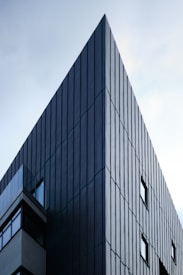 Angular modern building with dark vertical cladding panels, featuring multiple windows and a sharp edge extending into a light sky background.