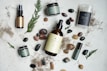 An arrangement of skincare or wellness products placed on a light surface, surrounded by natural elements such as leaves and assorted stones. The products include bottles with pump tops, jars, a spray, and a stick, each with minimalist labels in earthy tones. Dried plant material and small colorful pebbles add a rustic and organic touch.