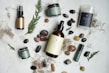An arrangement of skincare or wellness products placed on a light surface, surrounded by natural elements such as leaves and assorted stones. The products include bottles with pump tops, jars, a spray, and a stick, each with minimalist labels in earthy tones. Dried plant material and small colorful pebbles add a rustic and organic touch.