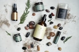 An arrangement of skincare or wellness products placed on a light surface, surrounded by natural elements such as leaves and assorted stones. The products include bottles with pump tops, jars, a spray, and a stick, each with minimalist labels in earthy tones. Dried plant material and small colorful pebbles add a rustic and organic touch.