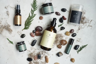 An arrangement of skincare or wellness products placed on a light surface, surrounded by natural elements such as leaves and assorted stones. The products include bottles with pump tops, jars, a spray, and a stick, each with minimalist labels in earthy tones. Dried plant material and small colorful pebbles add a rustic and organic touch.